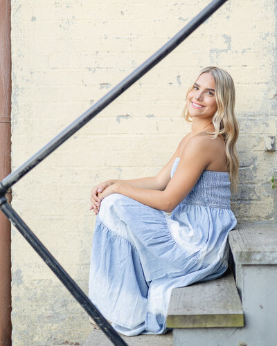 High school senior female with blonde hair, brown eyes, sitting on stairs against a yellow wall, smiling at the camera
