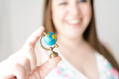Pinks & Greens Travel Co. CEO Rachel Salley holds  a miniature globe between her thumb and index finger out in front of her face. The globe is in focus, while her smiling face is out of focus in the background of the picture.