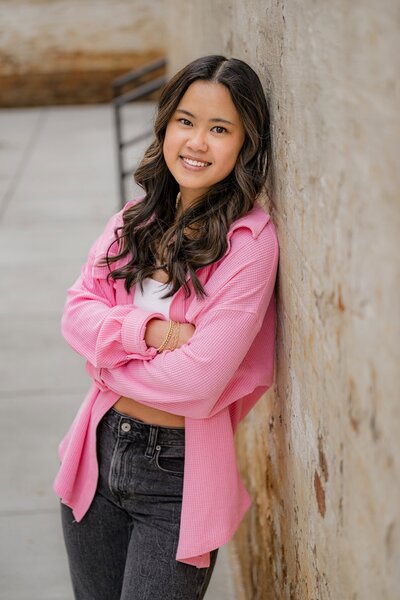 dark hair graduate leans against a wall wearing a hot pink shirt