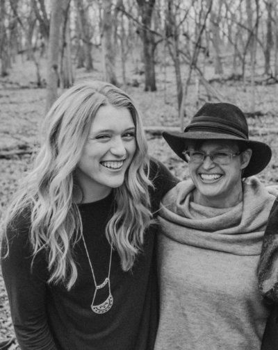 Mother and daughter laughing together and smiling at the camera. Photo is in black and white. Mother is wearing a hat and glasses and daughter is leaning in.