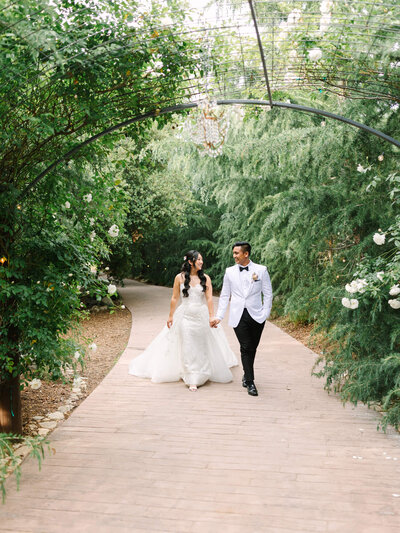 Bride in a white gown and groom in a white tuxedo walk hand in hand on a garden path, surrounded by lush greenery and arches.