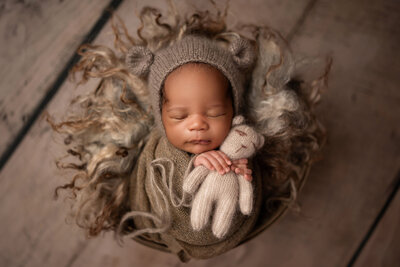 Newborn baby swaddled in earthy tones, wearing a knit bear bonnet and cuddling a matching teddy bear during a styled portrait session in Waukesha, Wisconsin