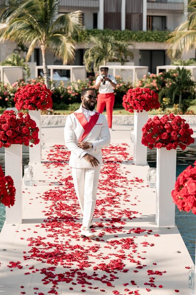 A man in a white suit walking down a dock with red petals.