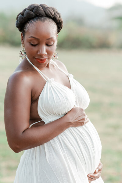 Pregnant woman of color standing  in a white dress holding her baby bump
