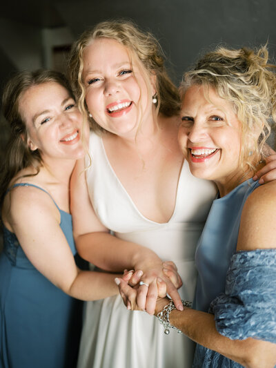 A bride, her mom, and her sister, hug, smiling at the camera in a portrait as she gets ready for her wedding.