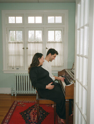 Couple from California in their home, playing the piano as the woman is in her black dress and holding her baby bump.