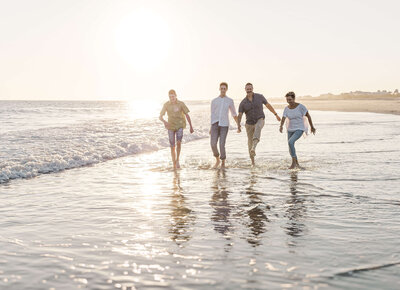 Four people holding hands walk along the shoreline of a sunny beach. The sun is low in the sky, creating a warm glow on the water. Captured by a Fayetteville NC family photographer, waves gently lap at their feet as they enjoy the beach atmosphere together.