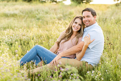 A white couple sitting closely in a grassy field, smiling at the camera.