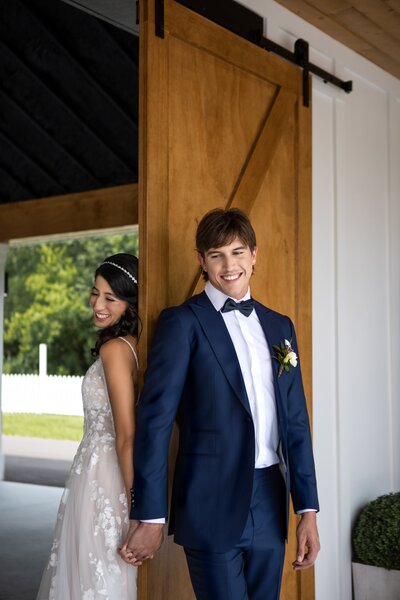 Bride and groom smiling as they lean against opposite sides of a door and hold hands