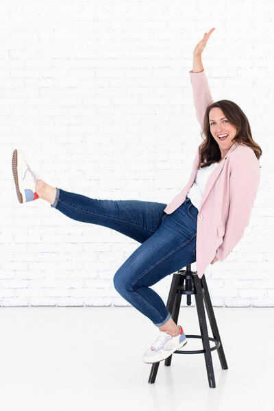 Social media strategist Katy is wearing pink blazer, white t-shirt and blue jeans. She is sitting on a small chair with no backrest and has her right arm and leg up as if she is playing. Behind her is a wall with white bricks and she is smiling at the camera.