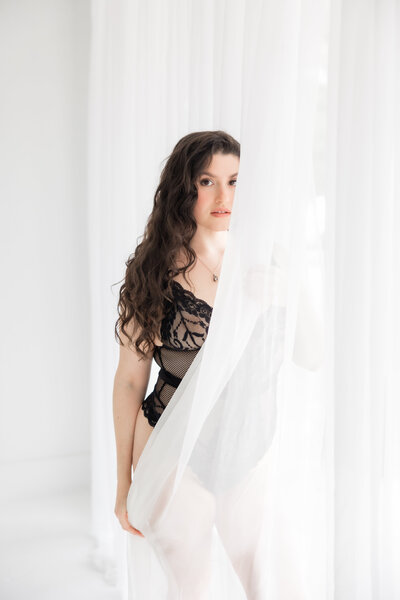 A woman in black lace behind a sheer curtain in a boudoir session at the Brand Studio Orlando