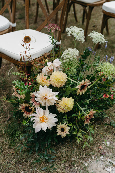 A lush garden-style wedding aisle floral arrangement featuring cream and pale yellow dahlias, soft blush blooms, Queen Anne’s lace, sunflowers, and layered greenery placed beside wooden cross-back ceremony chairs with white cushions, creating a natural, organic outdoor ceremony design.