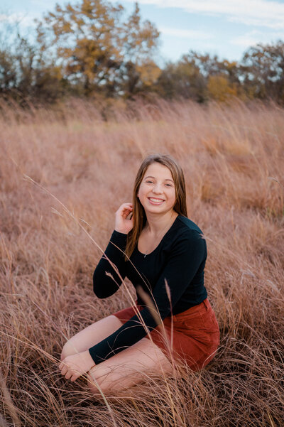 A high school senior poses for the camera kneeling in a field with one hand holding strands of her hair back.
