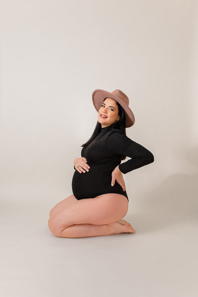 A mom-to-be in a black bodysuit and pink hat posing in a studio.