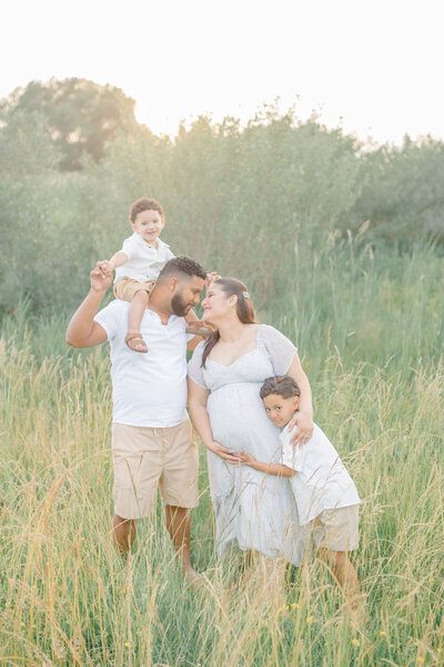 a family stands in a field with beautiful golden light, the mother holds her pregnant belly with their newest family addition. Captured by Portrait Studio Stamford CT photographer.