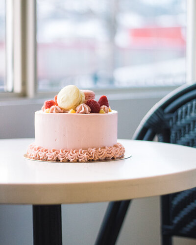 Elegant pink cake decorated with macarons, raspberries, and yellow fruit pieces on display at Gold Leaf Pastries in Kamloops BC.