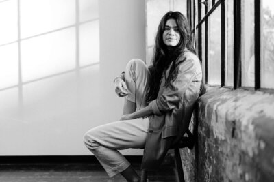 A person with long hair sits on a chair by a window, looking at the camera, in a black and white image, exuding the thoughtful elegance of a brand designer.