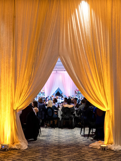 A luxurious event with people seated at tables beneath warm yellow drapes. The drapes are parted, framing a view of the room filled with guests, floral arrangements, and ambient lighting, creating an elegant and inviting atmosphere.