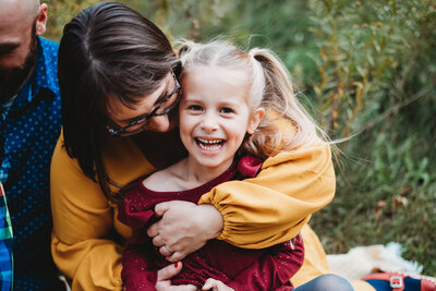 Little girl wearing red smiling at the camera as her Mom wraps her hands around her and hugs her in family photoshoot in Milwaukee, WI