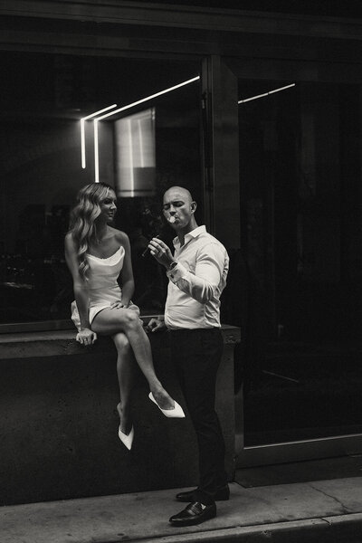 Urban elopement couple sharing a cigar outside a modern building at night, captured in black and white for a cinematic city feel.
