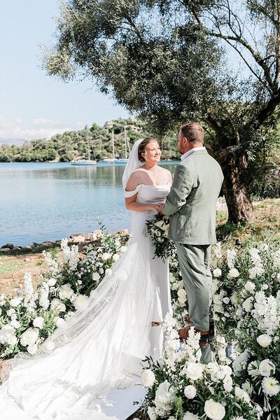 Bride and groom exchanging vows by the water under an olive tree, surrounded by lush white floral arrangements at their intimate destination wedding in Greece