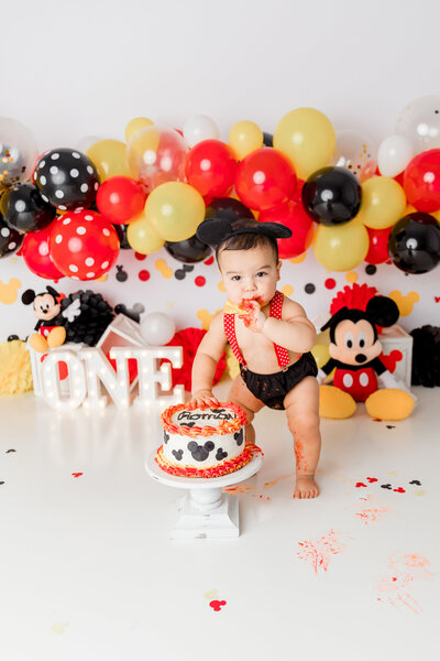 Adorable baby boy celebrating his 1st birthday with a Mickey Mouse-themed cake smash in a New Jersey photography studio. Dressed in red suspenders, black shorts, and Mickey ears, surrounded by vibrant red, yellow, and black balloons, and Disney-inspired decorations
