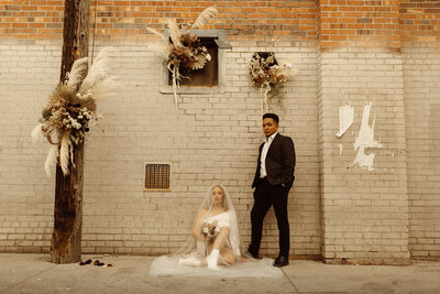 bride sitting and groom standing in front of brick wall styled with pampas grass and florals