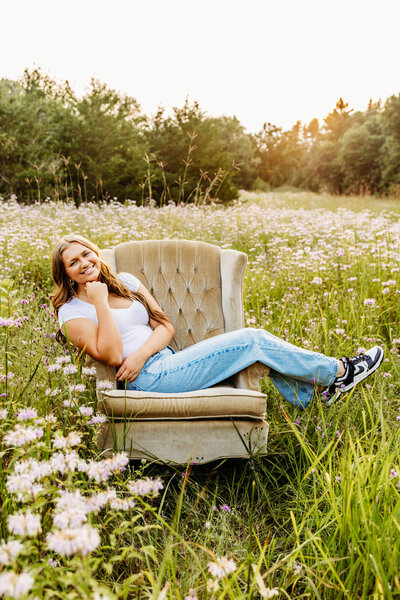 stunning teen girl in a white top and jeans sitting on a tan chair in a field full of flowers near Green Bay