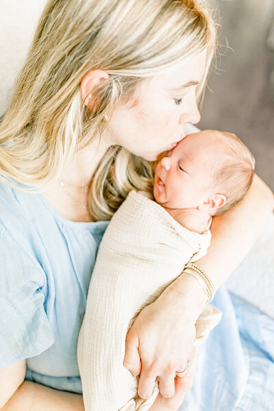 A newborn baby sleeps peacefully in her mother’s arms, dressed in a soft white outfit with a delicate headband. This light and airy portrait captures the tenderness of early motherhood, reflecting the warm and natural style of a Birmingham newborn photographer.