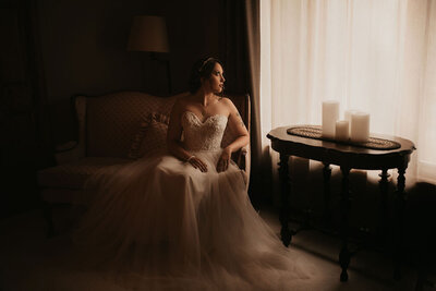A fine art portrait of a bride sitting on a vintage sofa in a flowing wedding dress. The room is dark, moody, and elegant. 