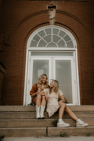 Photo of twin sisters sitting on the stairs of an old school looking at each other and laughing.