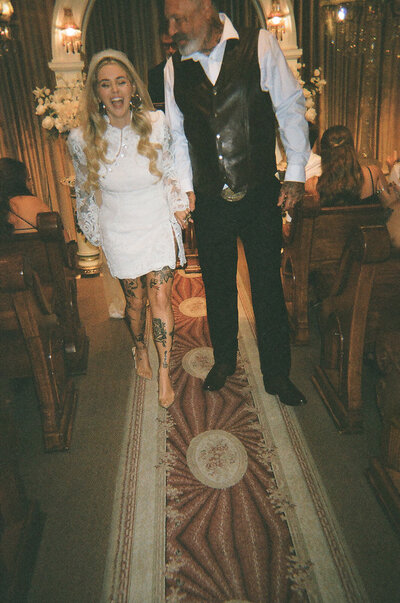 The couple from the previous image walking down the aisle of a chapel, both smiling and holding hands, with the bride in a short lace dress.