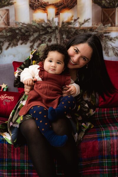 Mother sitting on a festive Christmas set with her baby on her lap, both dressed in warm holiday outfits and smiling for the camera.