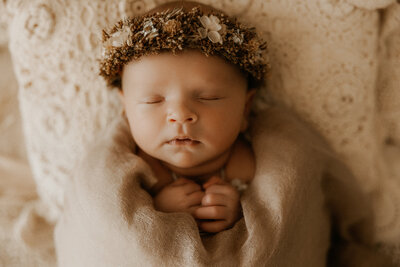 Photo of a newborn baby girl in a pretty newborn wrap leaning up against a pillow with a flower crown in the studio.