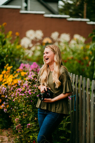 Staci laughing while holding a camera near a rustic wooden fence surrounded by wildflowers