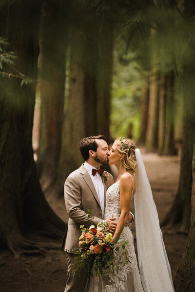 Calgary wedding photographer captures bride and groom sharing a kiss in a wooded area. Her dress is a stunning lace, and her wedding bouquet is full of muted red and pink flowers.