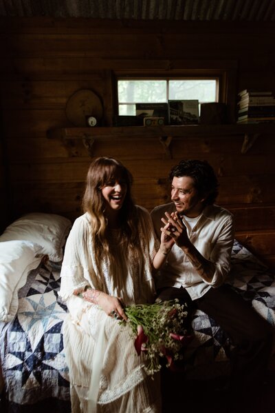 bride and groom smiling sitting on a bed holding hands and her bouquet