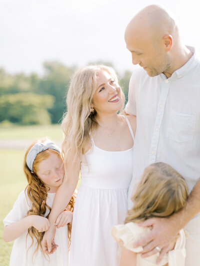 Blonde woman in white dress looks up at her husband while their two young daughters snuggle them at Little Rock park