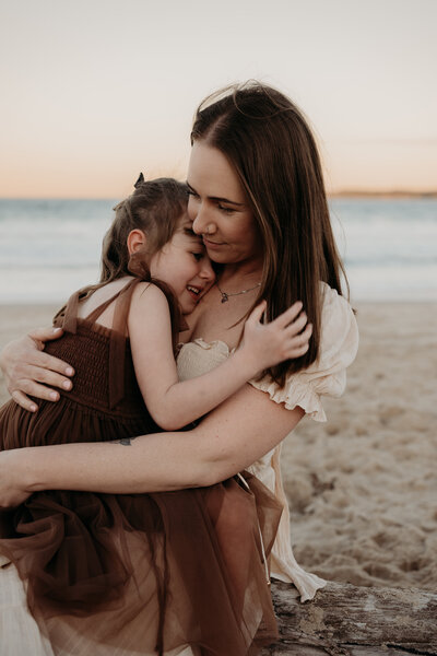 A woman and a young girl share a warm embrace on a sandy beach at sunset, perfectly capturing the essence of an NSW mother and child photography session.