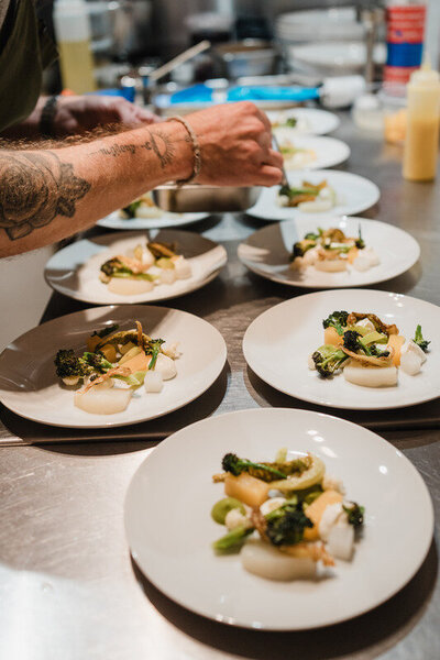 Chef plating vegetable dishes featuring broccoli, melon, and turnip on white plates in the kitchen.