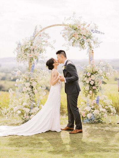 A bride and groom kiss under a floral arch during their outdoor wedding ceremony, surrounded by pastel-colored flowers and greenery – a picture-perfect moment made possible through meticulous wedding planning in Canada.