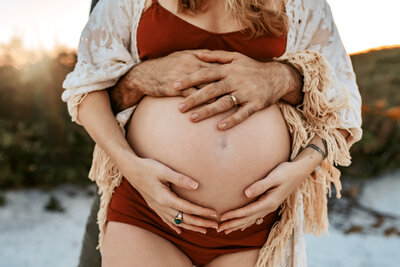 Maternity Photographer, a pregnant woman holds her belly and her husbands hands also fondly holds her belly as he stands behind her