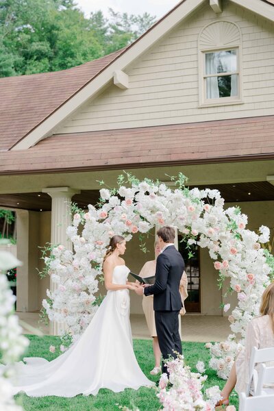 Beautiful wedding ceremony at Winbrooke in Brookshires, Massachusetts, featuring a stunning floral arch as the backdrop. The bride and groom exchange vows in an intimate outdoor ceremony, surrounded by lush greenery. Felisha, a destination wedding photographer based in New England, captures timeless moments at weddings in Massachusetts, New England, and worldwide locations.