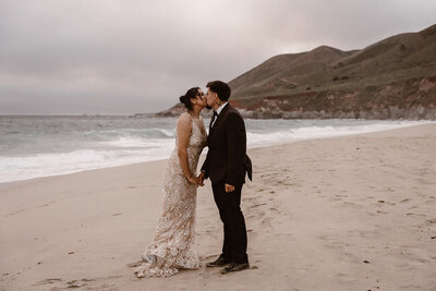 A couple shares a kiss on a sandy beach with ocean waves and coastal hills in the background.
