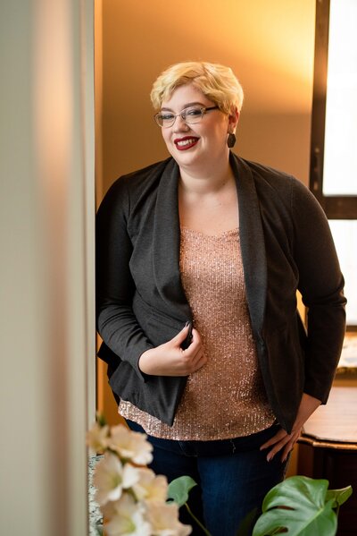 st-louis-maternity-photographer-irene-kinghorn-poses-against-wall-smiling-wearing-pink-sparkly-top-and-black-blazer