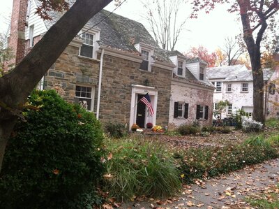 Historic stone home with an overgrown front yard, scattered fallen leaves, and an unkempt walkway leading to the entrance, with an American flag hanging by the door.