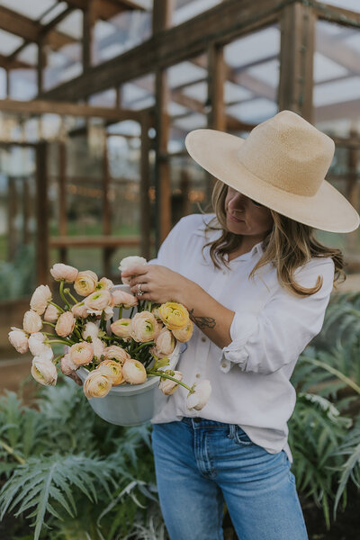 Owner of Peaches and Poppies Floral picking flowers for a bucket wearing a white button down shirt and large sun hat