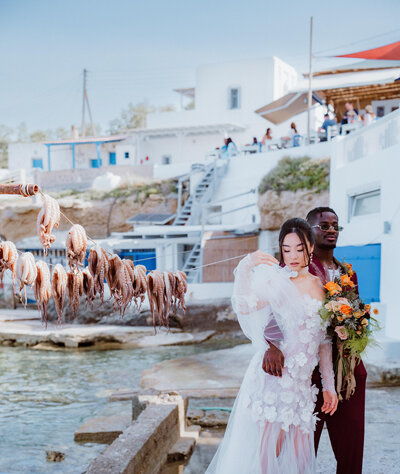 Bride and groom pose at a seaside Greek village with octopus drying on a line, showcasing a multicultural wedding with modern floral details and coastal charm.