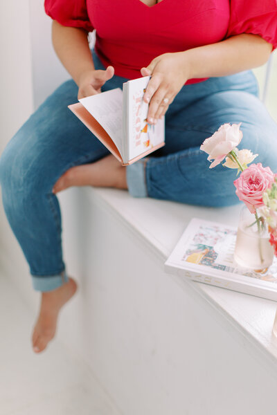 Woman sitting in a window sill wearing a pink shirt flipping through a book about cocktails
