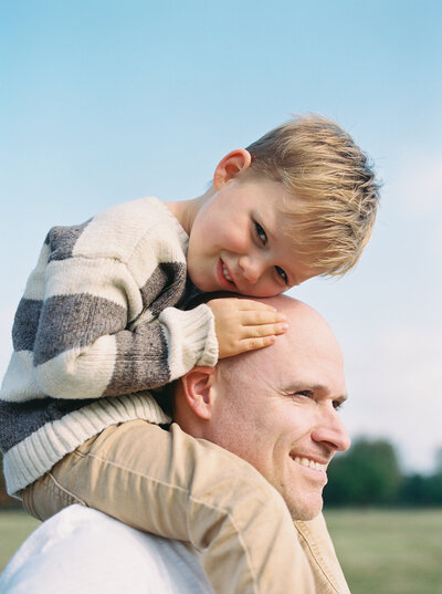 Film image of a boy smiling while rest his head on his father's head, as he sits on dad's shoulders